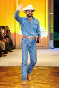 Model on runway wearing light blue denim outfit with a cowboy hat, walking confidently during fashion show