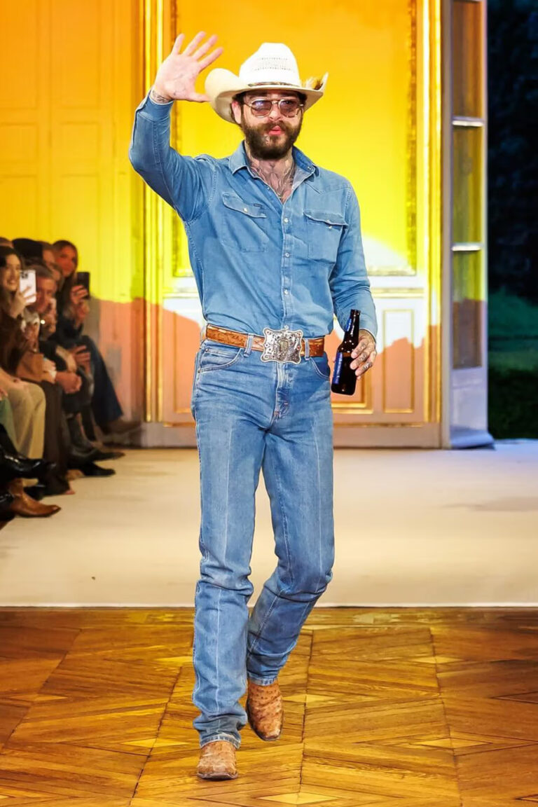 Model on runway wearing light blue denim outfit with a cowboy hat, walking confidently during fashion show